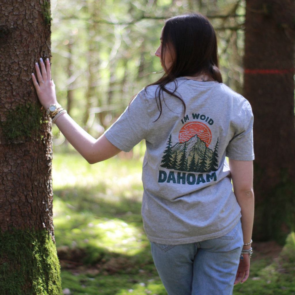 Nice Wave T-Shirt ‚Im Woid dahoam‘ in grau aus 100% Bio-Baumwolle. Nachhaltige Streetwear aus dem Bayerischen Wald mit bayerischem Dialekt-Spruch für Naturverliebte und Freigeister. Im Woid Dahoam frau trägt es im wald 